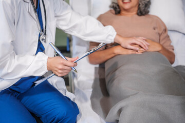 Caucasian female specialist doctor attends to an elderly Asian female patient in a hospital bed, providing medical care and support.