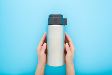 Young woman hands holding and showing closed new silver steel thermos with dark black plastic mug for hot drink or soup on pastel blue table background. Closeup. Point of view shot. Top down view. - 801203210