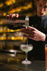 Fototapeta na wymiar Barkeeper pours creamy white drink through sieve into glass. Skilled young bartender prepares alcoholic cocktail for serving clients