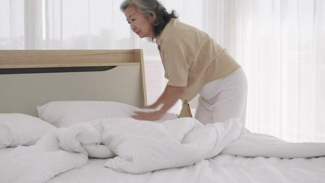 Footage Handheld medium shot, Asian elderly woman was cleaning the dusty bed in bedroom, She use hand closed her nose during arranges pillows and white duvet