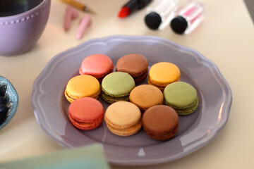 Plate of pastel macarons, cookies and chocolate, cup of tea of coffee, glass of bubble water, various berries, books and accessories on the table. Selective focus, pastel colors.