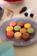 Plate of pastel macarons, cookies and chocolate, cup of tea of coffee, glass of bubble water, various berries, books and accessories on the table. Selective focus, pastel colors.