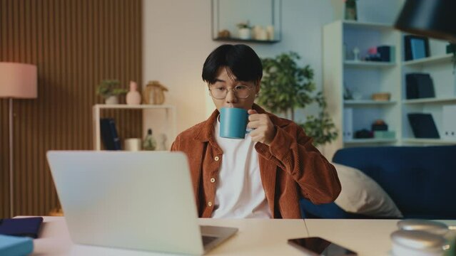 Relaxed Asian man in glasses opens laptop and drinks coffee, freelancer's day