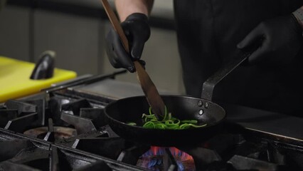 Close up a professional chef in a black uniform fries green vegetables and adds white pieces of seafood to a hot frying pan on a gas burner while preparing a dish in a restaurant kitchen