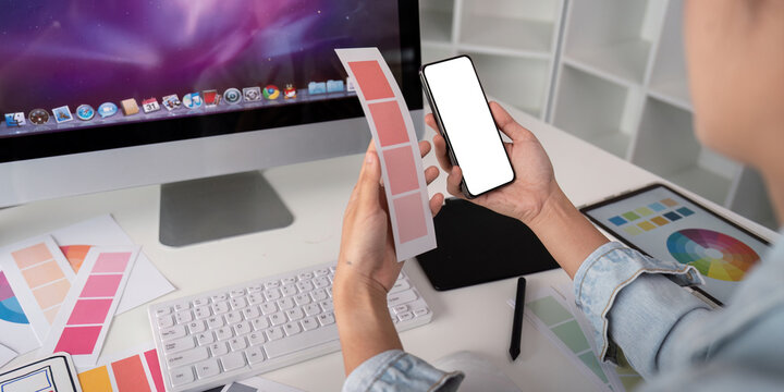 Woman hands holding a smartphone white screen mockup over modern graphic designer office desk