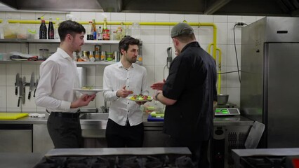 Two chefs in black uniforms and waiters communicate about serving and specific dishes in the kitchen in a restaurant. Workers in the kitchen of a restaurant communicate about the quality of serving