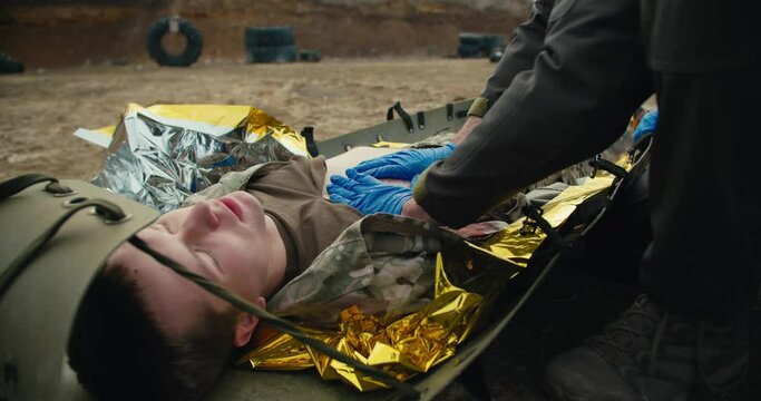 Close-up providing first aid to wounded soldiers on the battlefield under the supervision of a military medic. Confident male medic in blue rubber medical gloves stops the blood of a wounded soldier