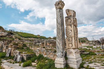 Intricate carved columns stand against the ruins of Aphrodisias, under a blue sky with fluffy...