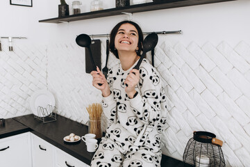 Cheerful woman standing in bright kitchen smiling laughing dressed in patterned pajama set. The...