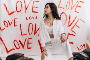 Fototapeta premium Woman sitting with kisses lipstick near black foil balloons, the word LOVE hand-painted on plain white surface oilcloth, Valentine`s day concept celebration decoration. Love, holiday, party 