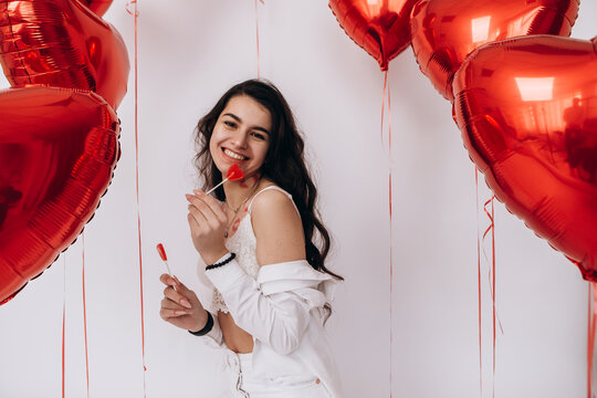 A smiling woman with kisses lipstick is holding eat candies and standing amidst heart-shaped helium balloons, celebrating a special holiday Valentines Day


