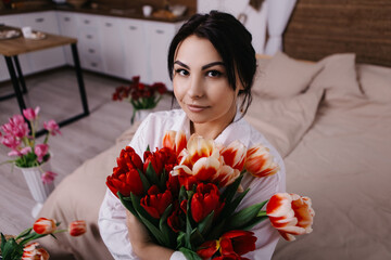 A woman poses on bed, surrounded by vibrant fresh bouquets bunch of red tulips placed around the room, domestic atmosphere. Spring International Women's Day, 8th of March. Holiday, festive 
