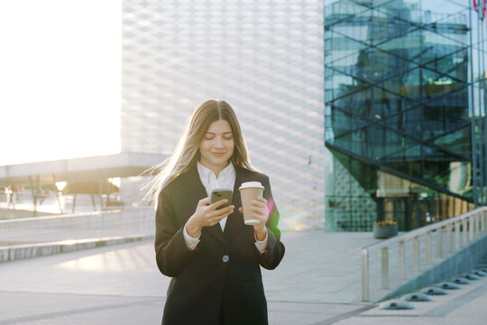 A businesswoman in a black suit walks with a coffee cup in front of office buildings looking at her phone
