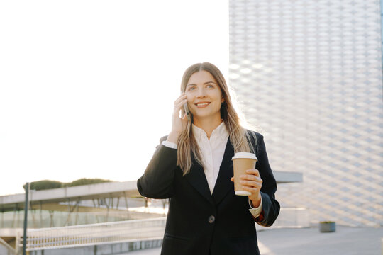 A smiling businesswoman in a black suit walks with a coffee cup in front of office buildings and talking on the phone