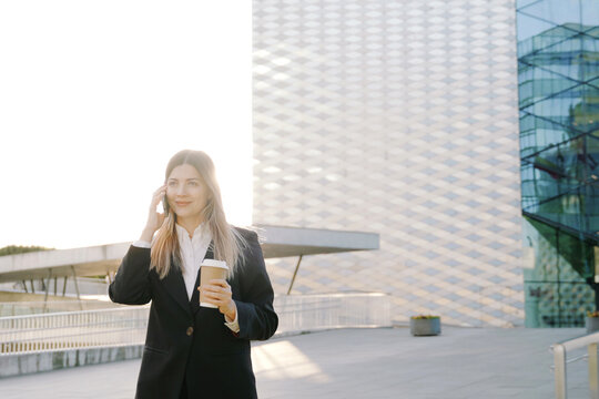 A smiling businesswoman in a black suit walks with a coffee cup in front of office buildings and talking on the phone
