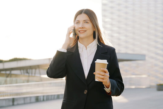 A smiling businesswoman in a black suit walks with a coffee cup in front of office buildings and talking on the phone
