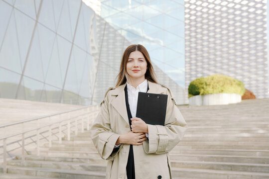 Confident young businesswoman in a white shirt and coat with a folder in her hands in front of office buildings