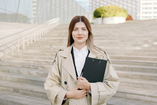 A portrait of confident young businesswoman in a white shirt and coat with a folder in her hands in front of office buildings