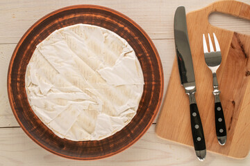Brie cheese on a clay plate with a fork and knife on a wooden table, macro, top view.