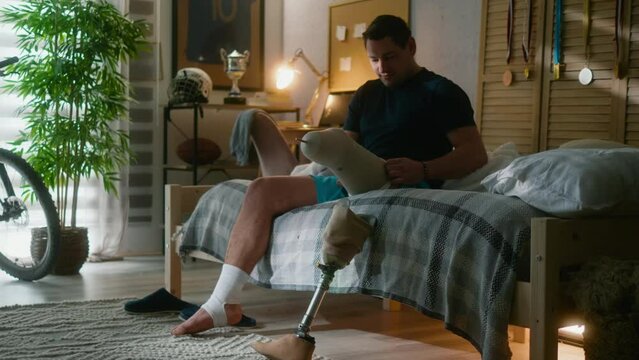 Young amputee man putting on prosthesis leg sitting on the bed early in the morning