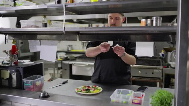 A confident male cook in a black uniform stands in the kitchen while issuing orders lays out stickers with written dishes and gives out the required dish to the hall for the waiters to hand over to
