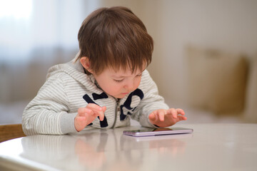 Pensive baby plays with a mobile phone while sitting at the table. The child looks at the phone screen. Kid aged two years