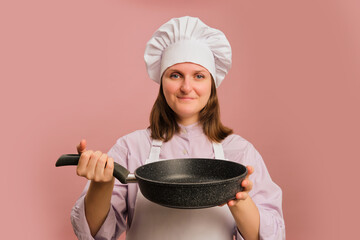 Woman chef holding frying pan on studio pink background. Portrait of a female person in chef's clothing