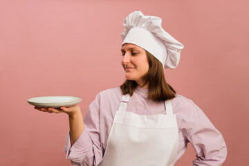 Woman chef holding empty saucer on studio pink background. Portrait of a female person in chef's clothing