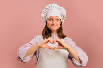 Woman cook showing heart with hands on studio pink background. Portrait of a female person in chef's clothing