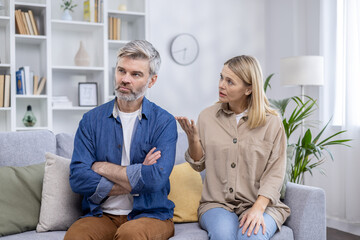 A mature man and woman having a serious conversation in a bright, modern living room. The man is...