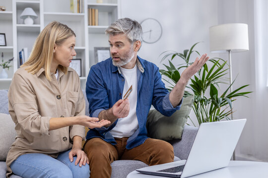 A mature couple engages in a heated discussion about online credit card fraud, gesturing at a laptop and credit card in a modern living room.