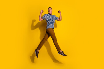Fototapeta na wymiar Full length photo of lucky funny guy wear print shirt jumping high rising fists emtpy space isolated yellow color background