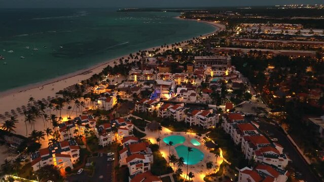 Aerial view of popular tourist coastal city at night. Luxurious tourist hotel with pool illuminated from below. Flickering lights of city below create mesmerizing picture. Punta Cana, Dominican