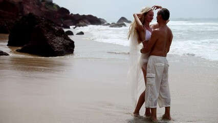 Groom gently hugs bride in swimsuit with long veil standing on sun kissed beach. Blonde fiancee adjusts wavy hair looking with smile at man