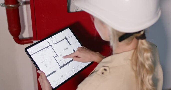 Woman engineer in helmet examines building plan on tablet. Attentive female worker checks fire safety standing near fire hydrant system