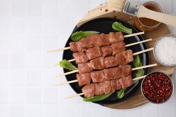 Wooden skewers with cut raw marinated meat , spices and basting brush on white tiled table, flat lay. Space for text