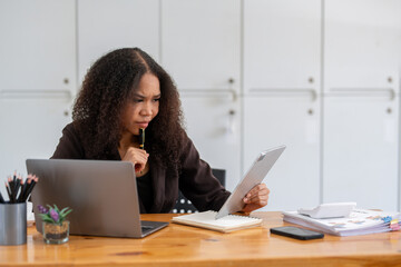 Stressed African American businesswoman reviewing documents while working on a laptop in a modern...