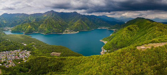 Panoramic aerial view of a lake surrounded by rugged mountains and forest (Saiko)