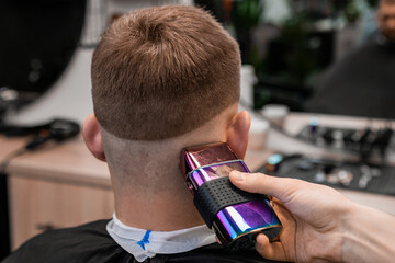 Hairstylist removes hair behind client ear and occiput with shaver in barbershop closeup. Barber uses modern machine for stylish haircut in salon