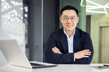A well-dressed Asian businessman smiles confidently while sitting at his modern office desk, portraying professionalism and success in a corporate environment.