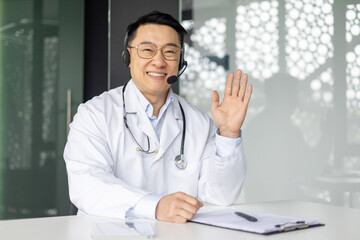 A cheerful Asian male doctor in a lab coat and headset waves hello during a telehealth consultation, sitting at his desk in a modern clinic. © Liubomir