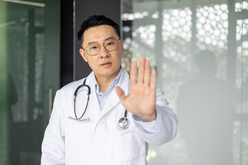 Confident Asian male doctor in a white coat with stethoscope, gesturing stop with his hand,...