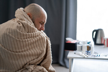 Bearded sick sad man wrapping blanket sits on couch suffers from flu disease. Unwell guy feeling...
