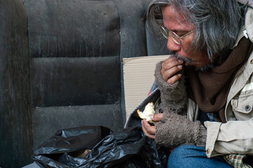 A homeless man sits on the sidewalk eating a piece of bread