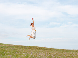 Quiberon, France, April 17, 2024. Woman with long red hair jumping against the blue sky, green grass nature. Happiness, freedom and physical fitness