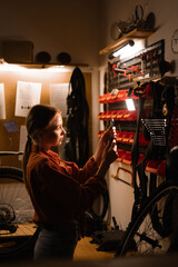 Obraz premium Female mechanic repairing bicycle in workshop, woman standing near wall with tools in garage. Bicycle maintenance concept.