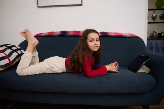 Caucasian Cute Elementary Age School Girl Smiling Looking At Camera, Lying On Sofa, Browsing Websites, Watching Movies And Having Fun On Her Digital Tablet With Black Mockup Screen.