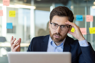 A middle-aged businessman looks confused and stressed while working on his laptop in a bright...