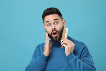 Surprised young man combing beard on light blue background