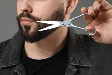Handsome young man trimming beard with scissors on grey background, closeup